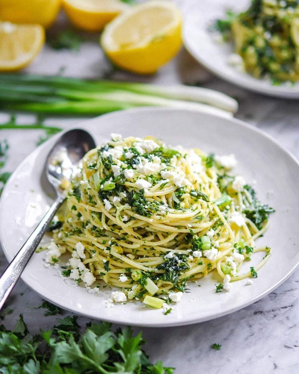 A white plate holds a mound of thin spaghetti pasta mixed with finely chopped green herbs and vegetables, giving the dish a fresh green tint with hints of yellow from the pasta. Crumbled white cheese is sprinkled over and around the pasta, adding texture and contrast. In the background, there is another white plate with more pasta and slices of bright yellow lemon. Fresh green herbs and spring onions are scattered on a white marbled surface surrounding the plate. A spoon is placed on the left side of the plate. The overall setting is bright and fresh, showing a healthy and herb-filled pasta dish. photo taken with an iphone --ar 4:5 --v 7